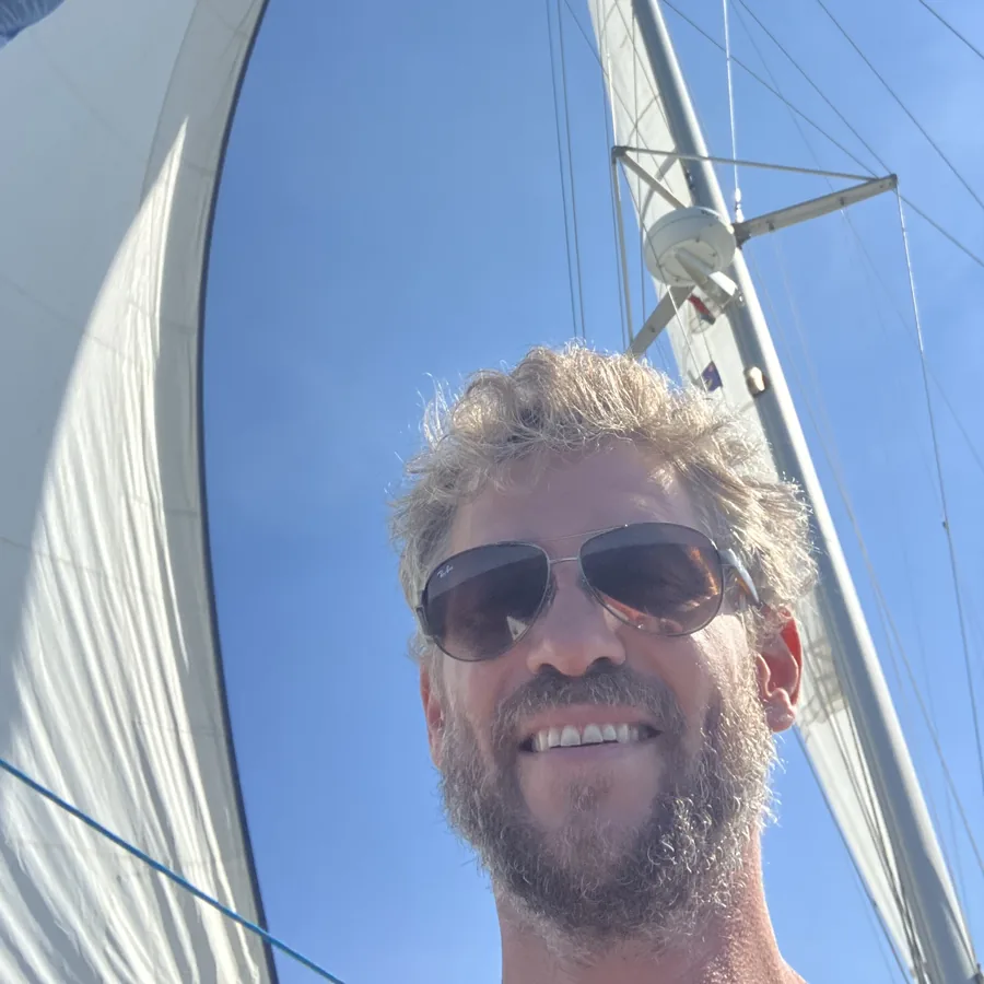 Thiash mit Sonnenbrille lächelnd an Bord eines Segelboots, hinter ihm Segel und Mast vor blauem Himmel